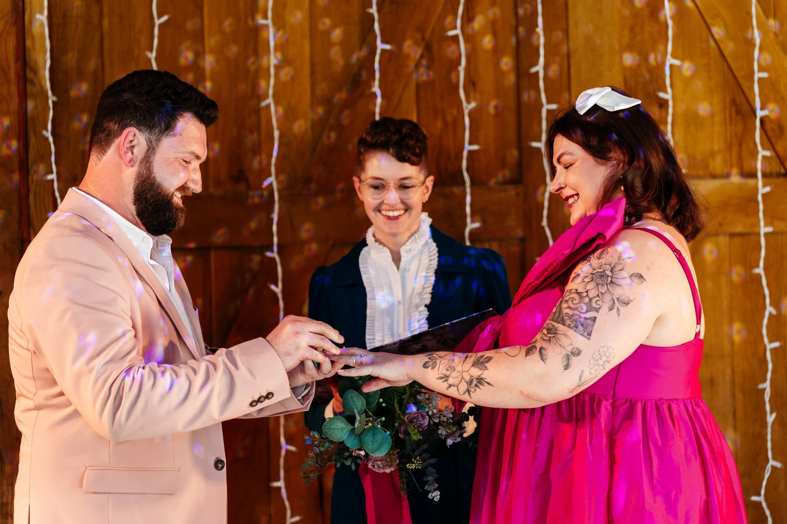 An indoor wedding ceremony in the UK featuring a smiling couple exchanging rings, with wedding celebrant Chloe Green officiating. The groom wears a light peach suit while the bride stuns in a bold pink dress, surrounded by warm wooden tones and soft 