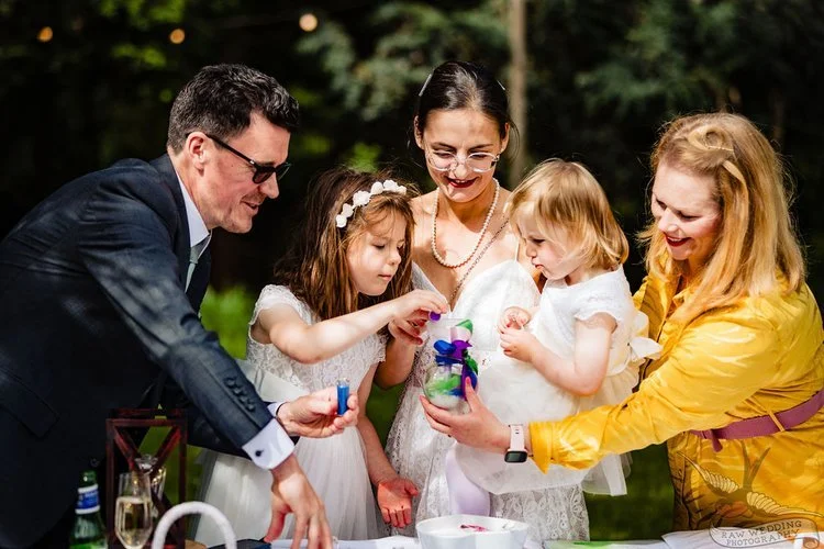 Outdoor wedding ceremony featuring a couple and two children participating in a colorful sand blending ritual. The bride and groom, along with their daughters, pour vibrant layers of sand into a glass jar, symbolizing unity and family. A joyful and h