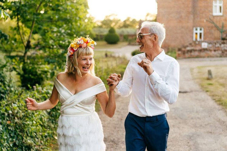 A couple share a joyful walk together in the evening sunshine, laughing and holding hands in a peaceful countryside setting, beautifully captured by Jordanna Marston Photography. The bride’s floral crown and modern off-shoulder dress paired with the 
