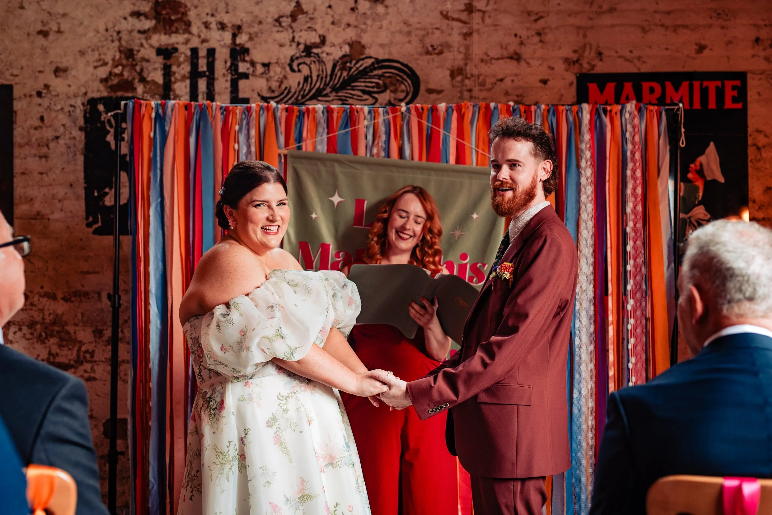 A joyful wedding ceremony led by Frances Celebrates You, an inclusive and fun UK wedding celebrant known for colourful, heartfelt celebrations. The couple stand laughing and holding hands in front of a bright, ribbon-filled backdrop while Frances del