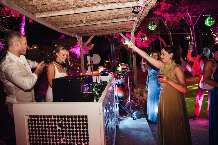 Mr and Mrs L performing at an outdoor reception while guests dance and celebrate under disco balls and colourful lighting. The lively setup blends professional sound equipment with a fun festival-style atmosphere, perfect for couples wanting high-ene