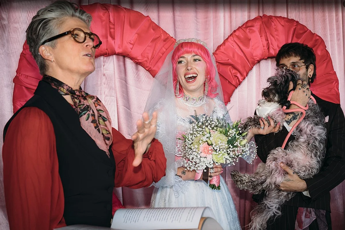Arabella stands to one side holding an open book, mid-gesture, while the couple face her beneath a large heart-shaped backdrop. One partner smiles broadly while holding a bouquet, the other cradles a small dog. Joanna Bongard Photography.