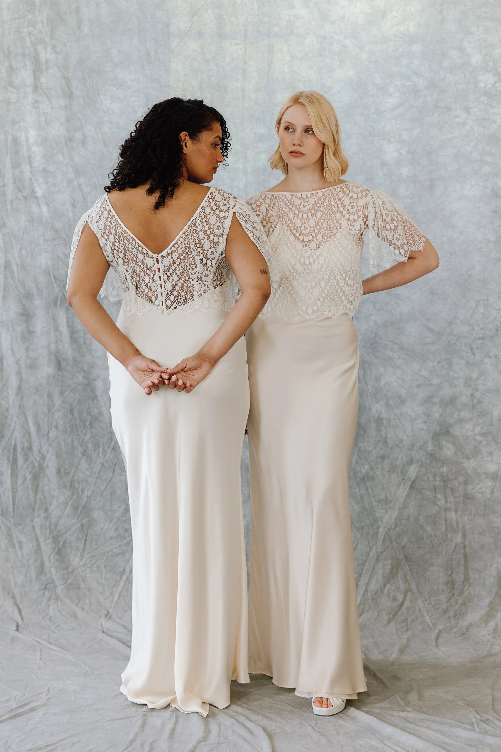 Two brides stand together against a soft textured backdrop wearing matching silk slip dresses topped with delicate lace blouses, one facing forward with hands on hips and the other turned slightly away with fingers loosely intertwined behind her back