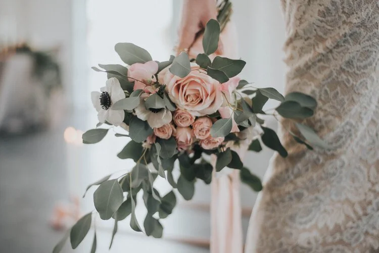 Romantic bridal bouquet by Catherine Short Florals featuring blush roses, white anemones and soft eucalyptus with flowing silk ribbons. Held against an elegant lace gown in a light filled setting, this delicate modern arrangement highlights Catherine