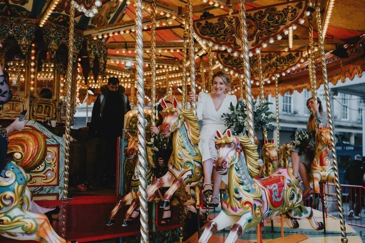 A playful and colourful wedding moment captured by Matt Fox Photography, showing the bride riding a beautifully decorated carousel horse while holding a lush bouquet of greenery. The warm glow of the carousel lights and the ornate painted details cre
