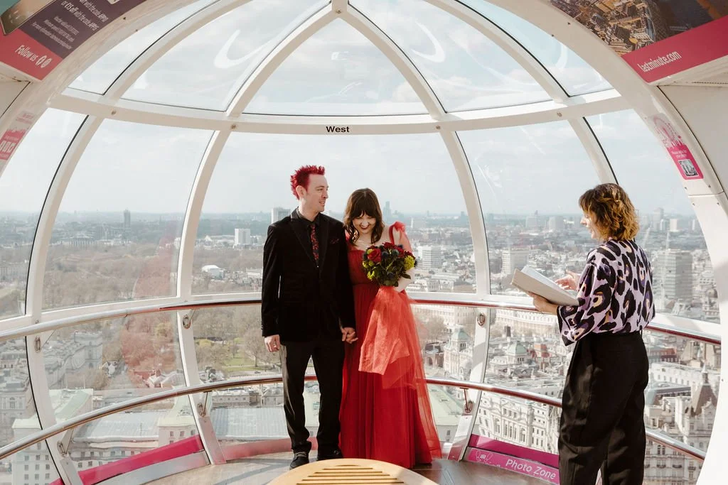 Cherry and Fern Celebrations leads a unique London wedding ceremony inside the iconic London Eye, with panoramic city views in the background. The couple, dressed in alternative wedding attire with the bride in a bold red gown, share a joyful moment.