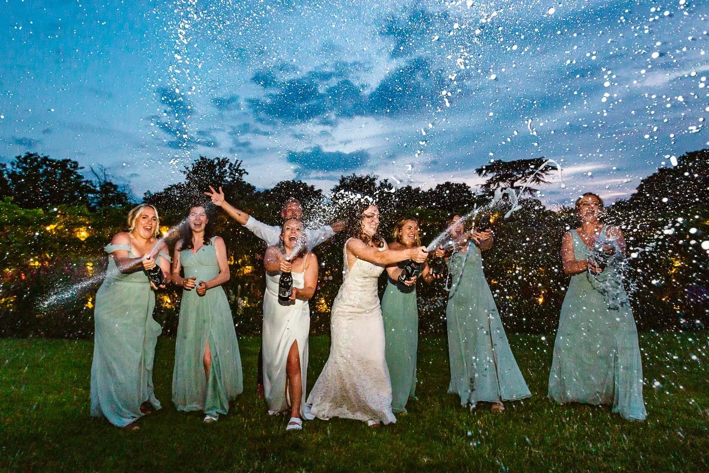 A joyful wedding party celebration captured outdoors at dusk, featuring a bride and her bridesmaids in matching light-green dresses popping champagne together. The group stands on a lush lawn as champagne sprays dramatically into the air, illuminated