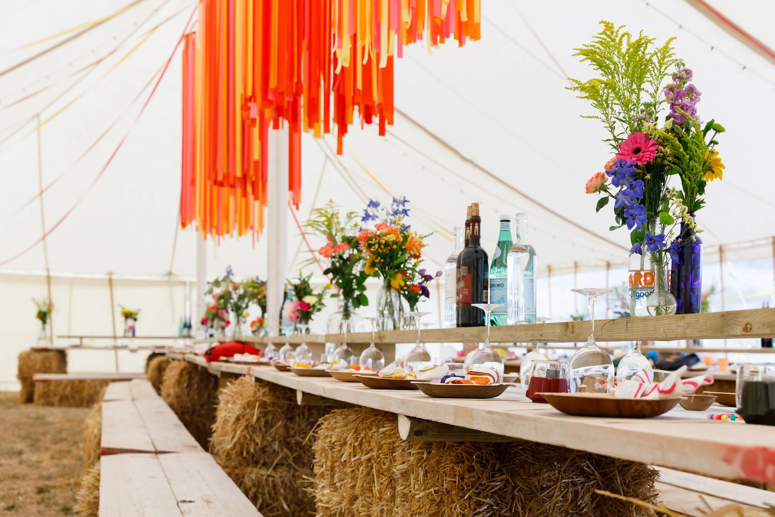 A colourful festival-style wedding reception setup inside a bright marquee, featuring long rustic trestle tables made from wooden planks and straw bale seating. The tables are decorated with vibrant, mismatched wildflower arrangements in reused bottl