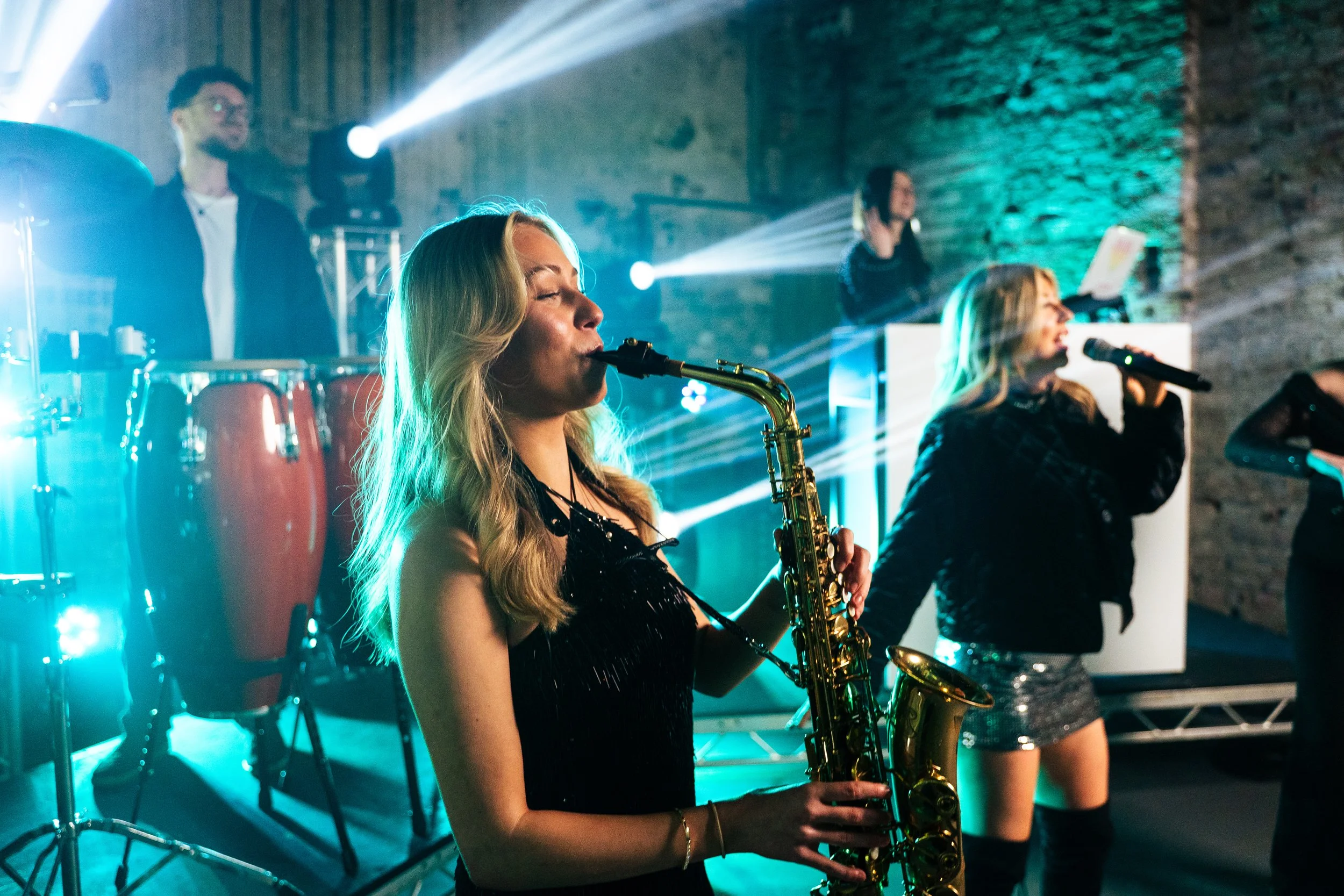 The Married to the Music wedding band performs live with saxophone, vocals, percussion, and DJ under vibrant teal and white lighting. This high-energy group brings modern wedding entertainment to life, creating an unforgettable party atmosphere for l