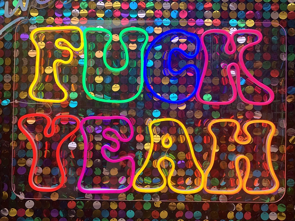 Vibrant neon wall art reading ‘F*ck Yeah’ in bold, colorful lettering against a shimmering rainbow sequin backdrop. This eye-catching, playful sign features glowing outlines in red, orange, yellow, green, blue, and pink, creating a dynamic and energe