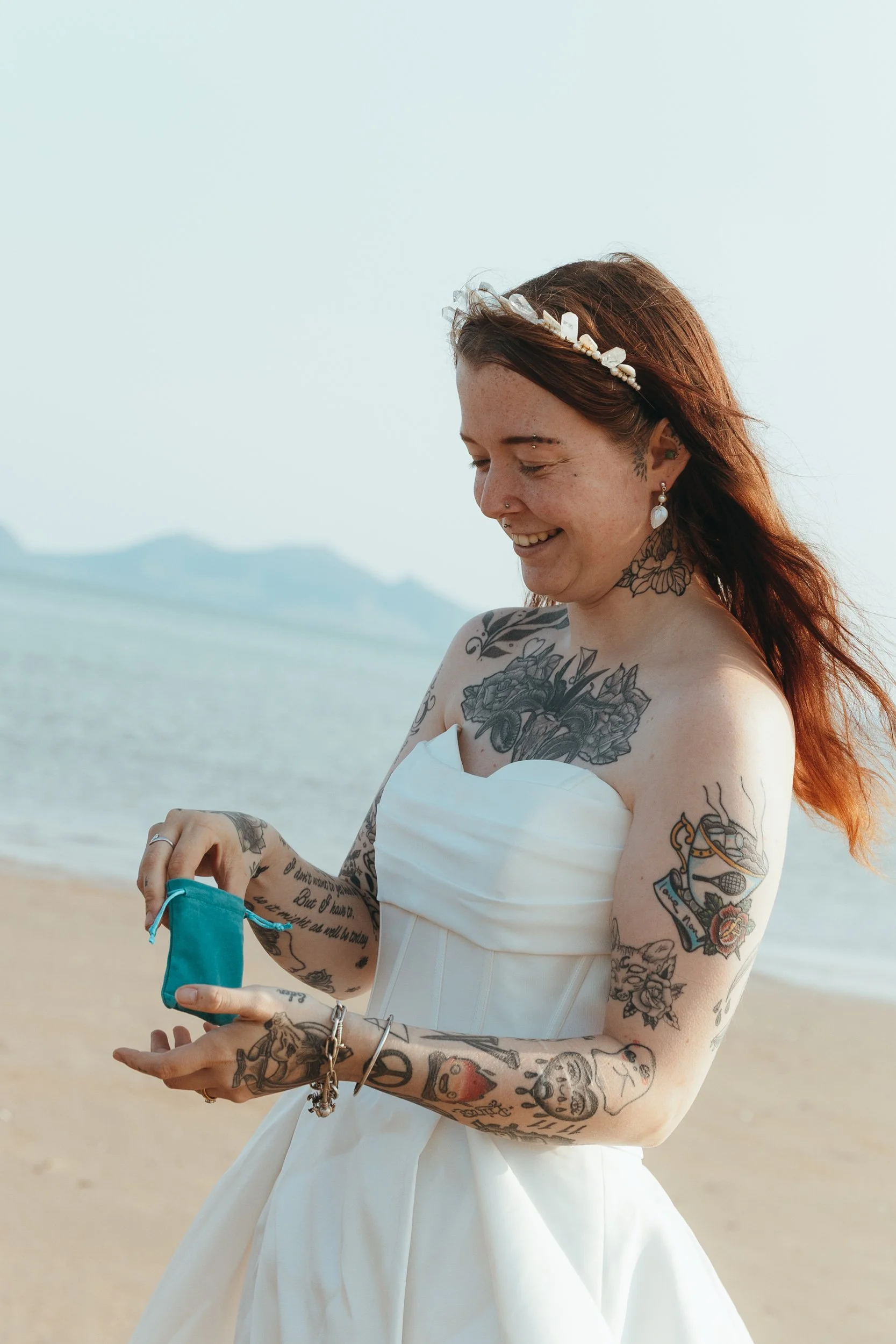 A Bride in a white strapless wedding dress smiling warmly, holding a small turquoise pouch. The backdrop features calm water and distant mountains, creating a serene and romantic atmosphere. Photography by Ariel in Blue.