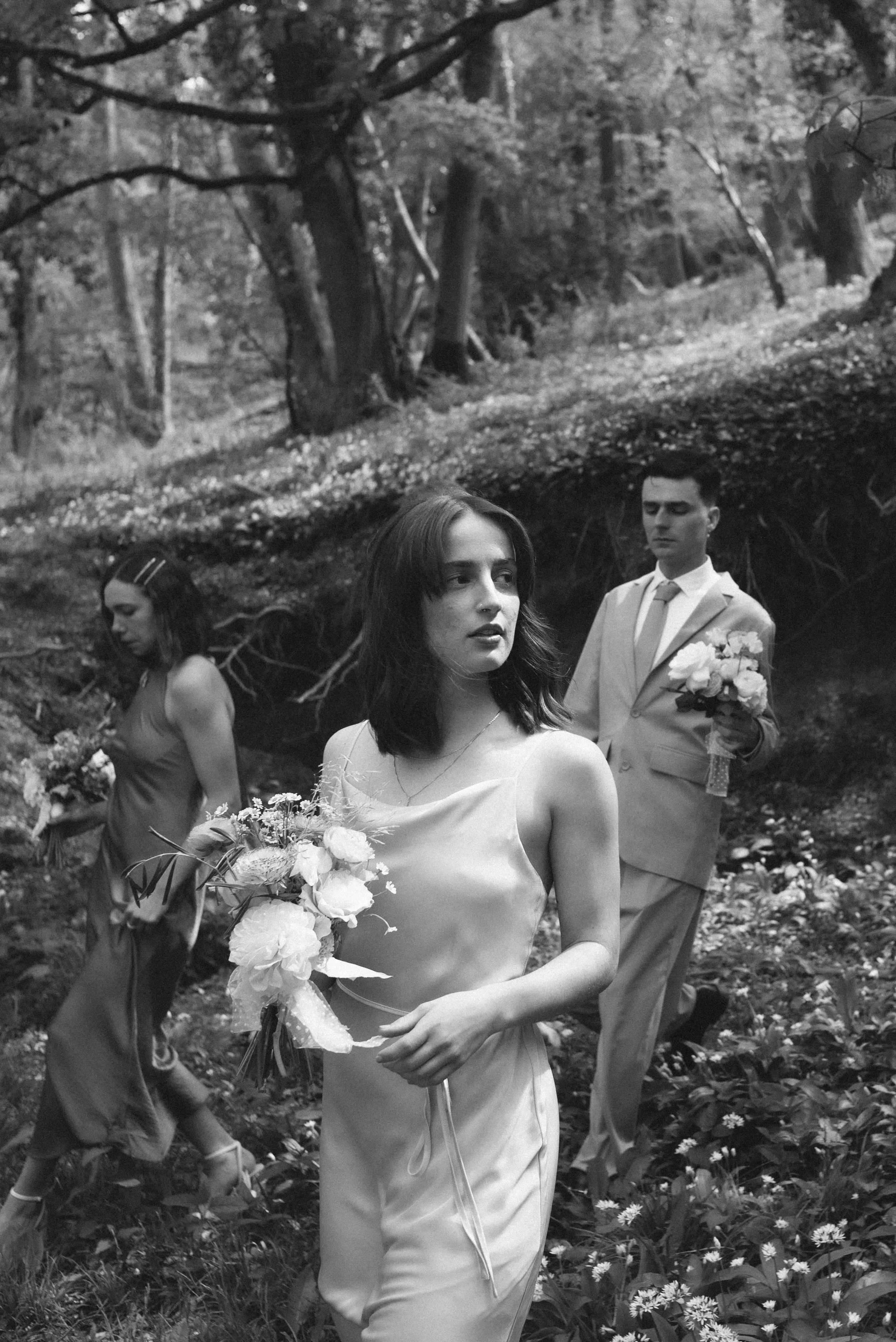Bridesmaids and a groomsman walk through a woodland clearing holding soft pastel bouquets, captured by Roz Pike Photography. The black and white documentary style and natural surroundings create an atmospheric editorial moment from a modern outdoor U