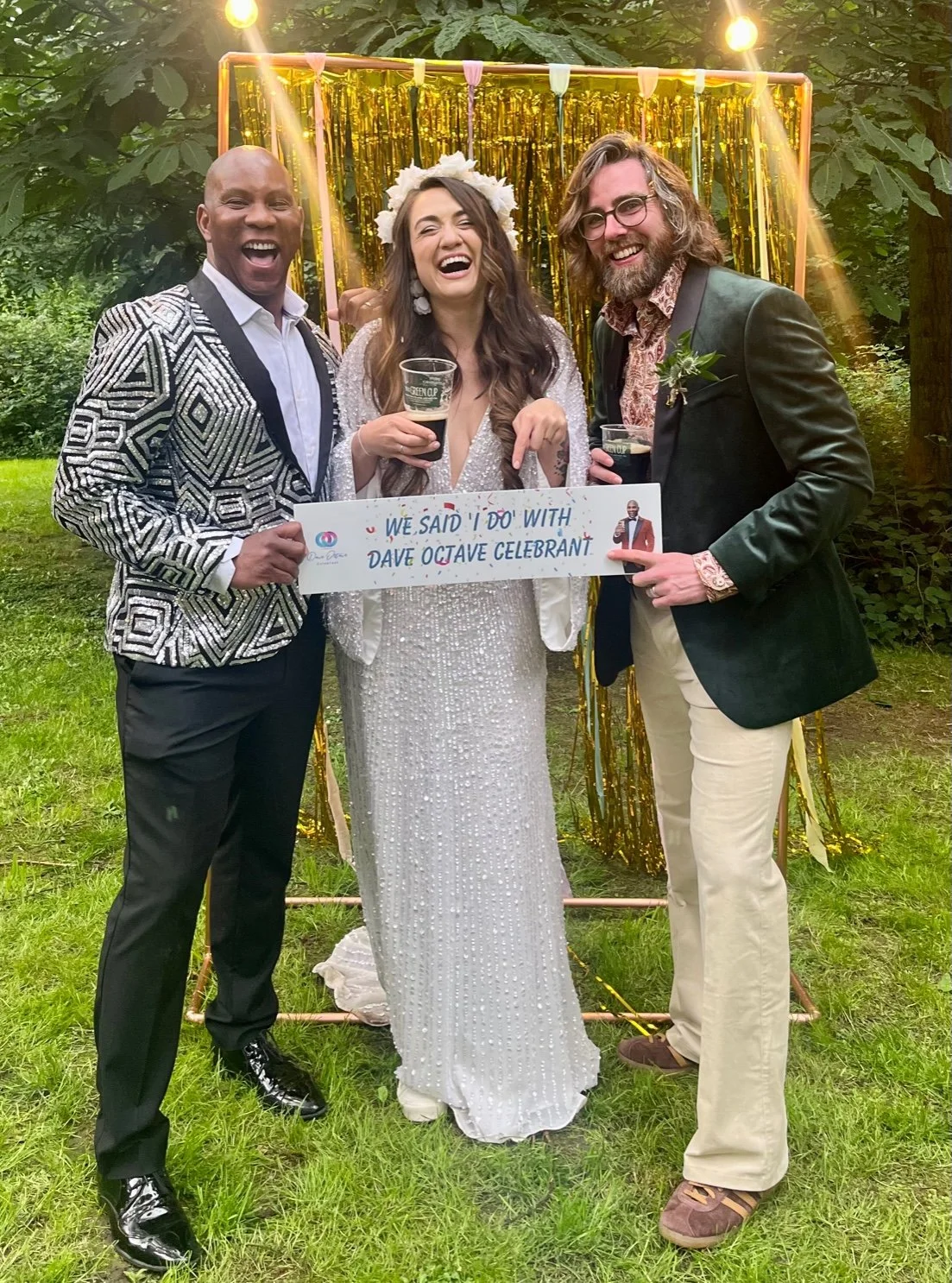 Dave Octave Celebrant in a black and white zebra print blazer, with a bride and groom, holding a sign that reads 'We said "I do" with Dave Octave Celebrant". 