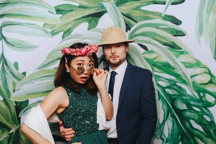 A couple strike a playful pose in front of a tropical leaf backdrop from Flash Fox Photo Booth. With fun props like sunglasses, a flower crown and safari hat, this booth captures the perfect mix of humour and style. Ideal for couples looking to add p