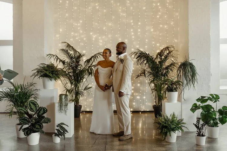 Modern minimalist wedding ceremony setup by Studio Clarisa Floristry featuring lush green plants in white pots and warm fairy light backdrop, creating an elegant natural atmosphere for the couple’s vows. Photos by Hash.
