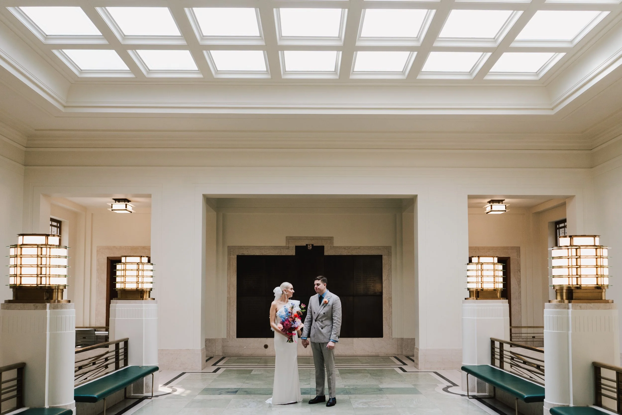 A couple stand together in the grand interior of Hackney Town Hall, framed by its striking Art Deco architecture, geometric skylights, and glowing vintage light fixtures. The bride’s sleek modern gown and vibrant, colourful bouquet contrast beautiful