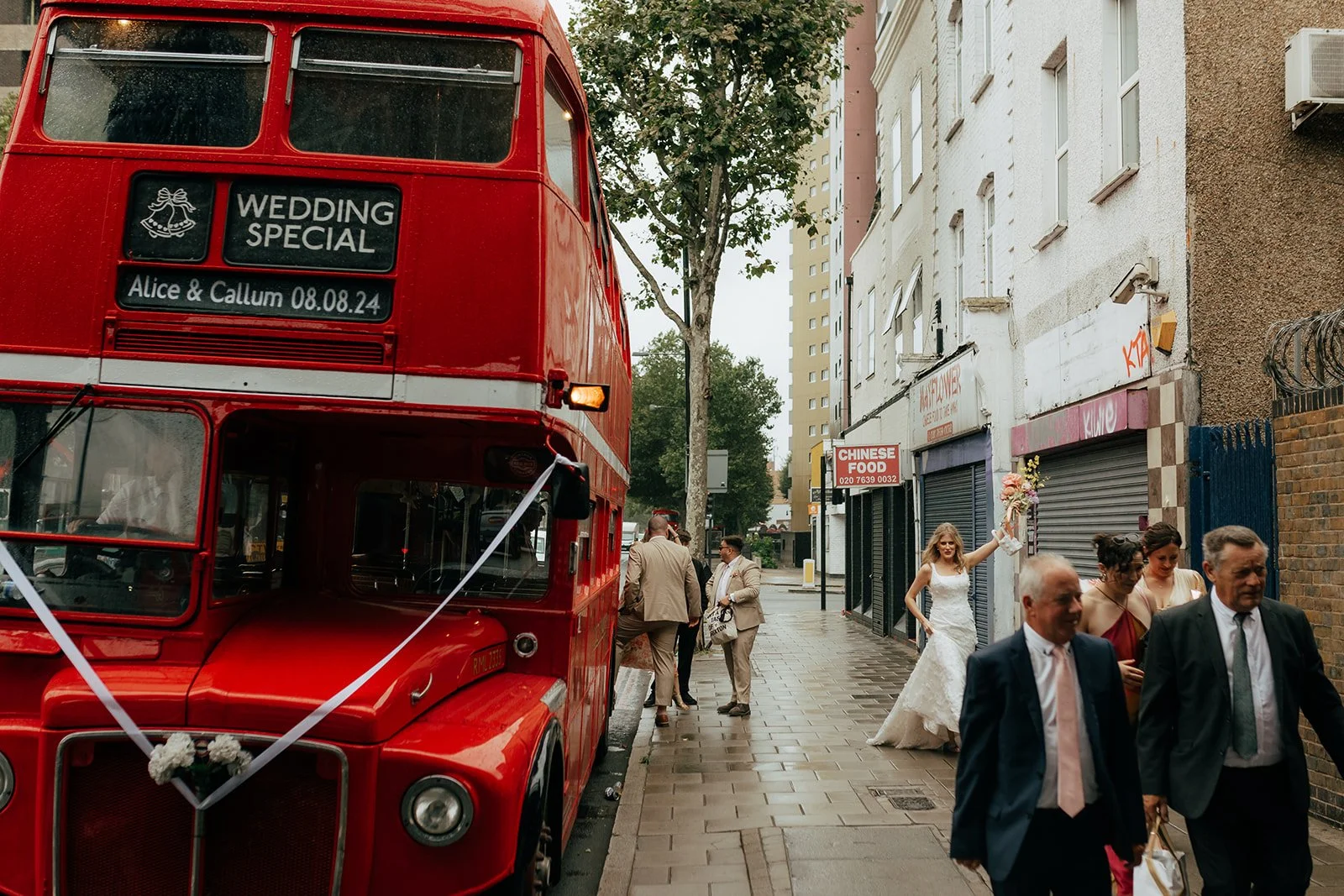 A stylish London wedding moment featuring a classic red Routemaster bus decorated with white ribbons and a personalised “Wedding Special” sign for Alice & Callum, 08.08.24. Guests in chic neutral outfits gather on the pavement while the bride, holdin