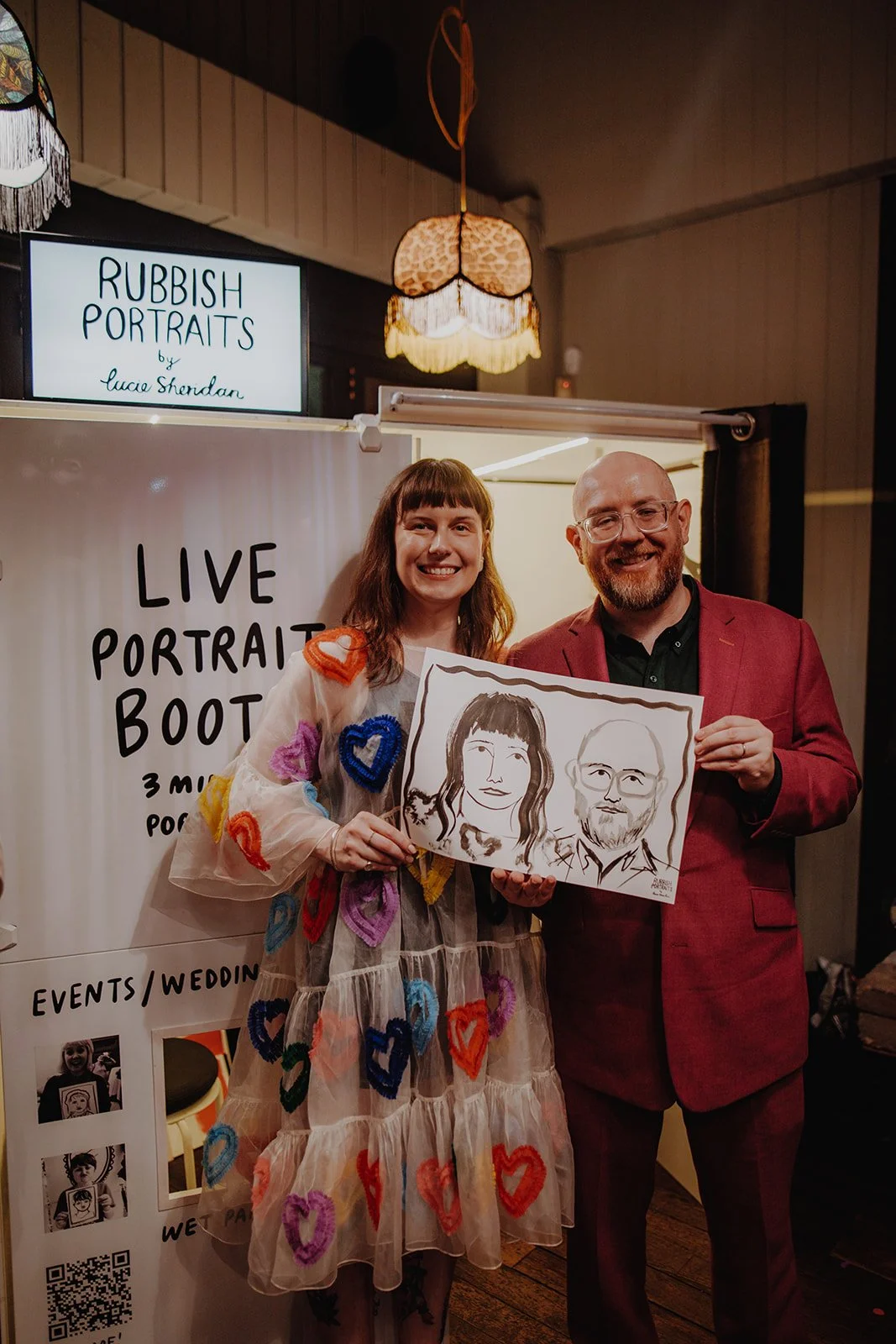 A happy couple stand proudly beside the Rubbish Portraits live portrait booth by Lucie Sheridan, showing off their hand-painted portrait. This quirky wedding entertainment idea brings creativity and laughter to any celebration, offering guests a uniq