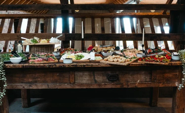 An impressive rustic buffet spread by Regis Banqueting, featuring a colourful array of gourmet dishes including fresh salads, meats, cheeses, and canapés. Set on a wooden table inside a barn-style venue, this elegant wedding catering display highligh