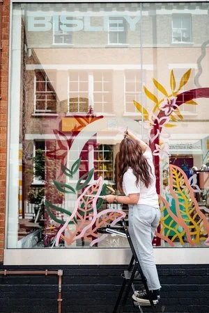 Artist Bee Davies painting a colourful botanical window mural for Bisley. Featuring bold hand-painted leaves and abstract shapes, this large-scale artwork showcases her signature illustrative style - perfect for brands and couples seeking creative, b