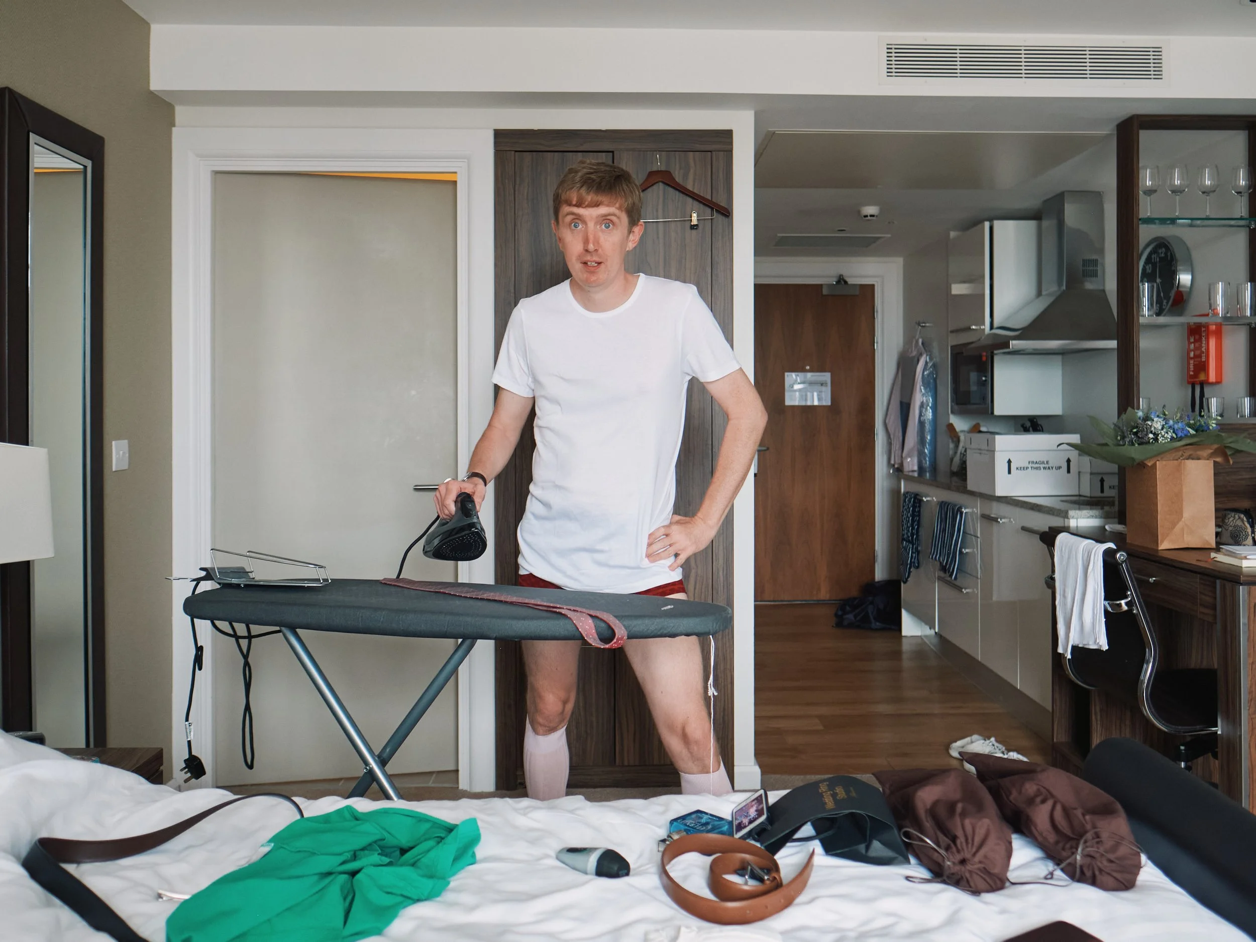 A candid and humorous getting ready moment captured by Wayne La Photography, showing the groom standing in the middle of a hotel room with an iron in hand as he prepares for the day. Dressed casually in a white T shirt, socks and boxers, he looks up 