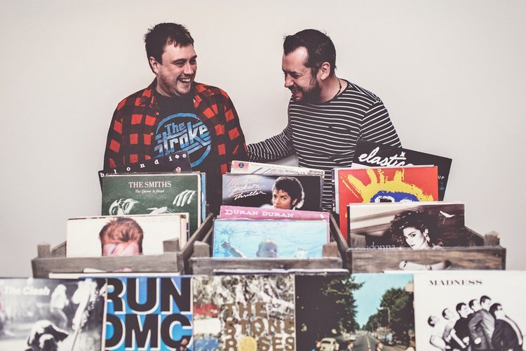 Two DJs from The Best Men stand behind stacks of classic vinyl records, smiling as they prepare their next wedding set. Surrounded by iconic albums from The Smiths, Duran Duran, and RUN DMC, they bring a love of great music and a fun, nostalgic vibe 