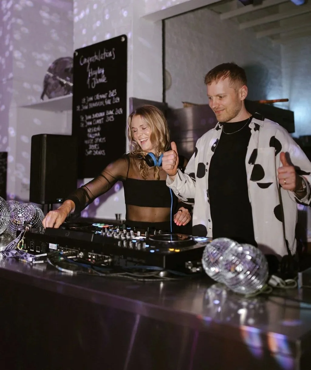 Mr and Mrs L are DJing together at a wedding. There are disco balls on the mixing desk.