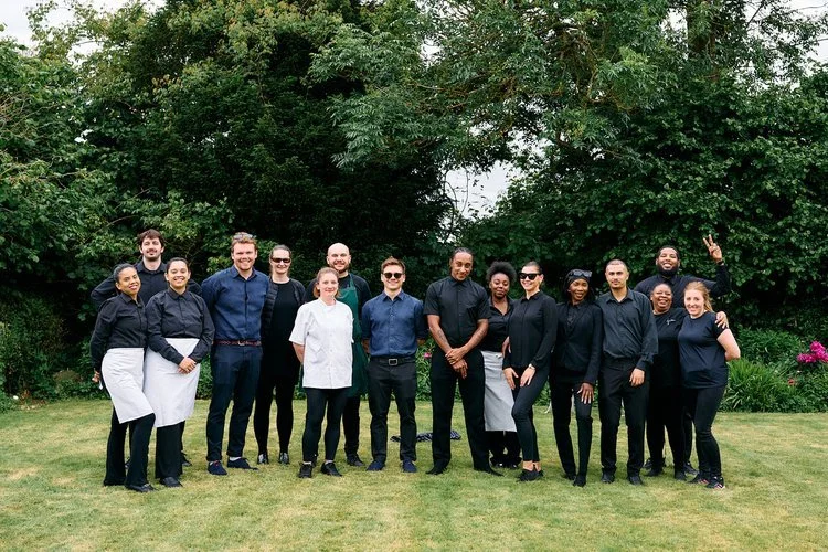 The professional catering team from Tasting Plates Wedding Caterers posing together outdoors before a wedding service. Dressed in smart black uniforms, the friendly staff stand on a manicured lawn surrounded by greenery, reflecting the company’s comm