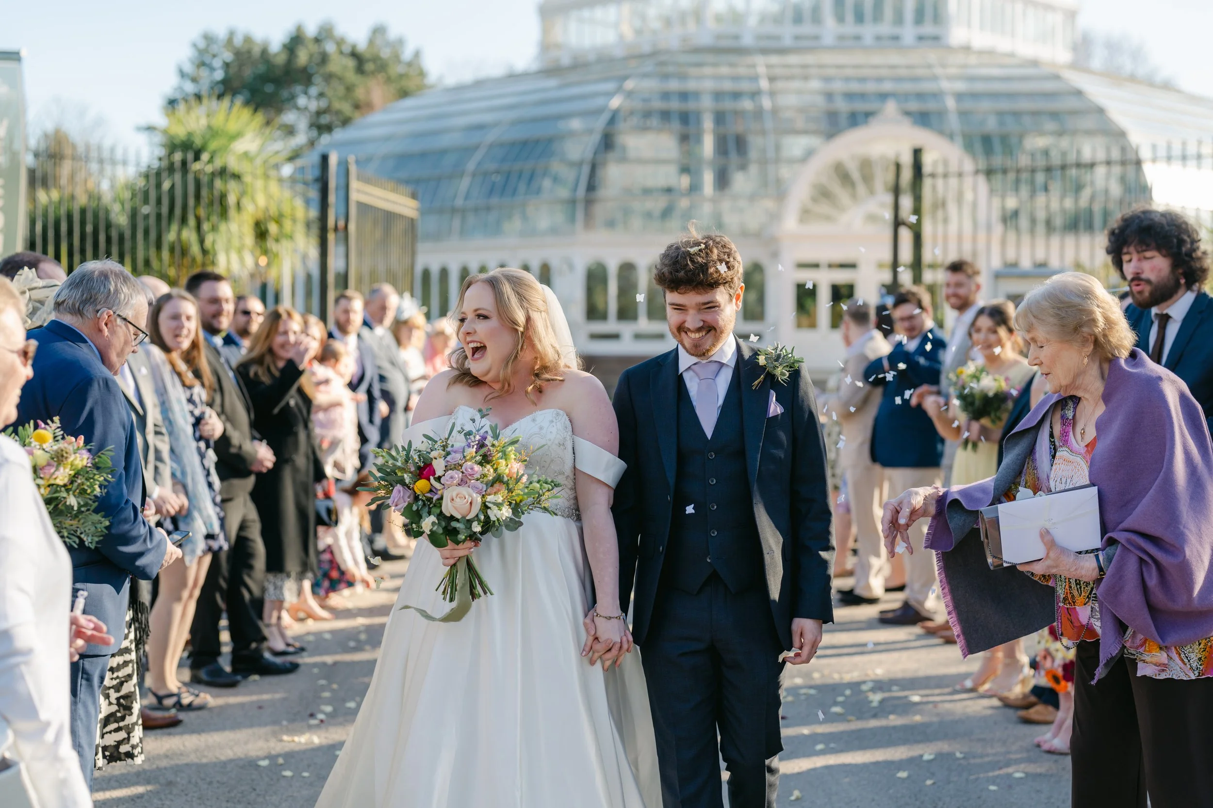 A newly married couple walk hand in hand through a joyful confetti tunnel outside a grand glasshouse venue, beautifully captured by Christy Photography. Surrounded by cheering guests and soft spring sunlight, the bride and groom share a moment of pur