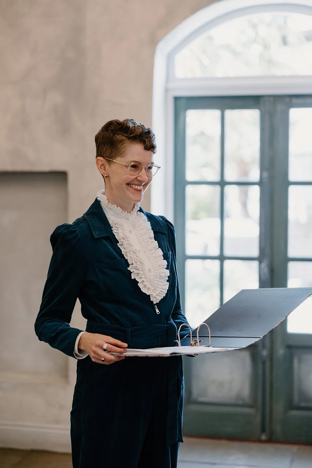 A smiling Chloe Green celebrant stands indoors holding an open ceremony folder, ready to officiate a modern and personal wedding ceremony.