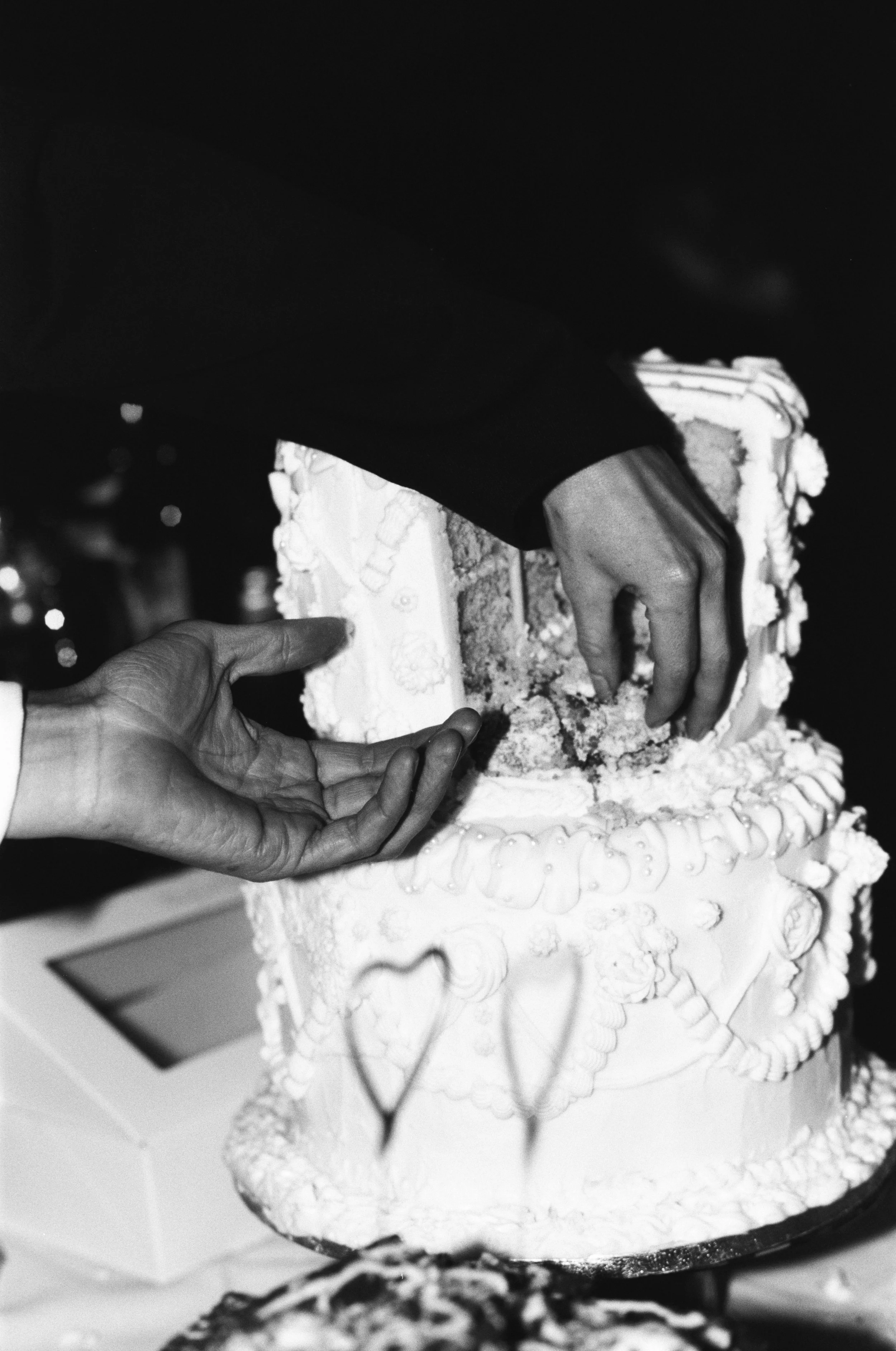 Hands reach into a vintage-style wedding cake as guests share slices during the celebration, captured by Roz Pike Photography. The black and white documentary style highlights the playful candid moment and the textured icing details of a relaxed mode