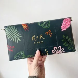 Hand-painted tropical clutch bag by Bee Davies Illustration, featuring personalised initials and wedding date in gold lettering. With bold botanical artwork and vibrant colours, this bespoke accessory is perfect for brides seeking unique, custom wedd