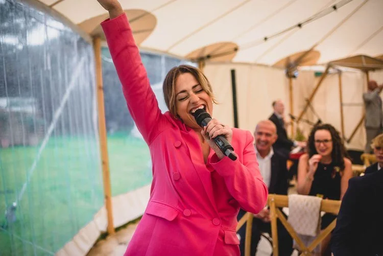 Wedding celebrant Vicky in a bright pink suit holding a microphone and leading a joyful moment during a wedding celebration.