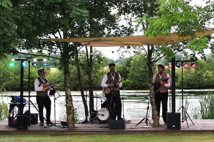 The Ukes of Hazard perform live on an outdoor stage beside a scenic river, surrounded by lush greenery. Dressed in their signature tweed waistcoats and smart shirts, the band members play ukuleles and sing beneath a canopy, creating a relaxed festiva
