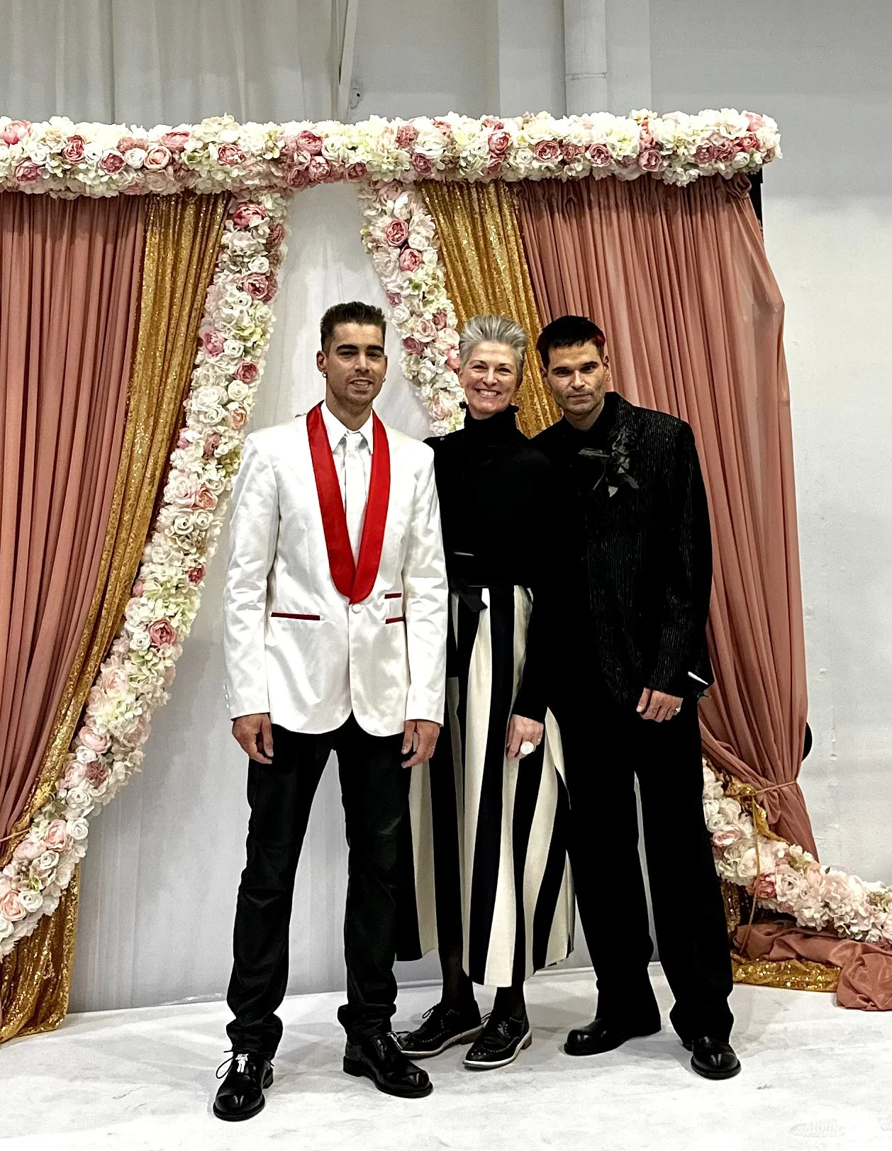 Three people stand together in front of a floral arch made from pale pink and cream flowers with soft pink draping and gold accents. The person on the left wears a white jacket with a red lapel detail, Arabella stands in the centre in a black top and