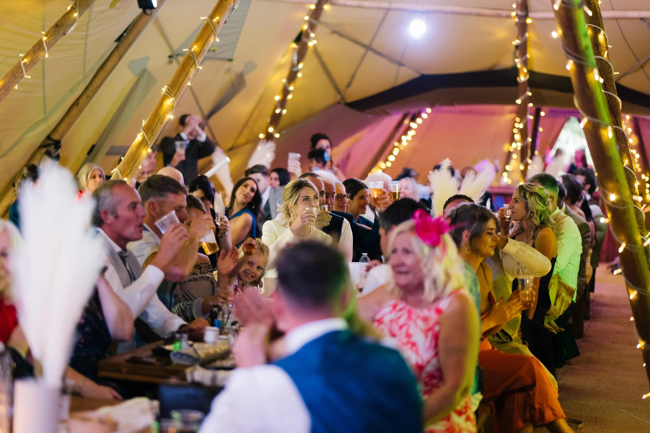 A warm lively scene unfolds inside a fairy lit tipi as guests sit shoulder to shoulder at long wooden tables lifting their drinks in a cheerful toast while soft golden lights wrap around the poles overhead casting a festive glow across smiling faces 