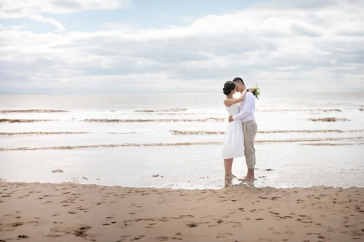 A couple share a quiet, romantic embrace at the water’s edge on a peaceful sandy beach, beautifully photographed by Christy Photography. Soft waves, open skies and natural light create a serene coastal backdrop that highlights the couple’s connection