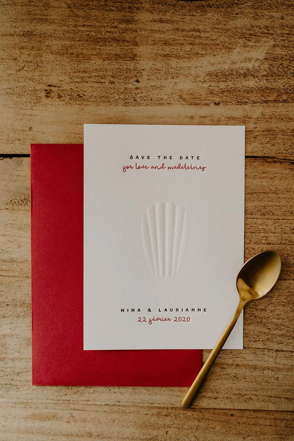 A minimalist wedding save the date flatlay featuring an elegant white card with an embossed shell motif, contemporary typography in black and red, a rich red envelope and a gold spoon arranged on a rustic wooden surface creating a refined fine art ae