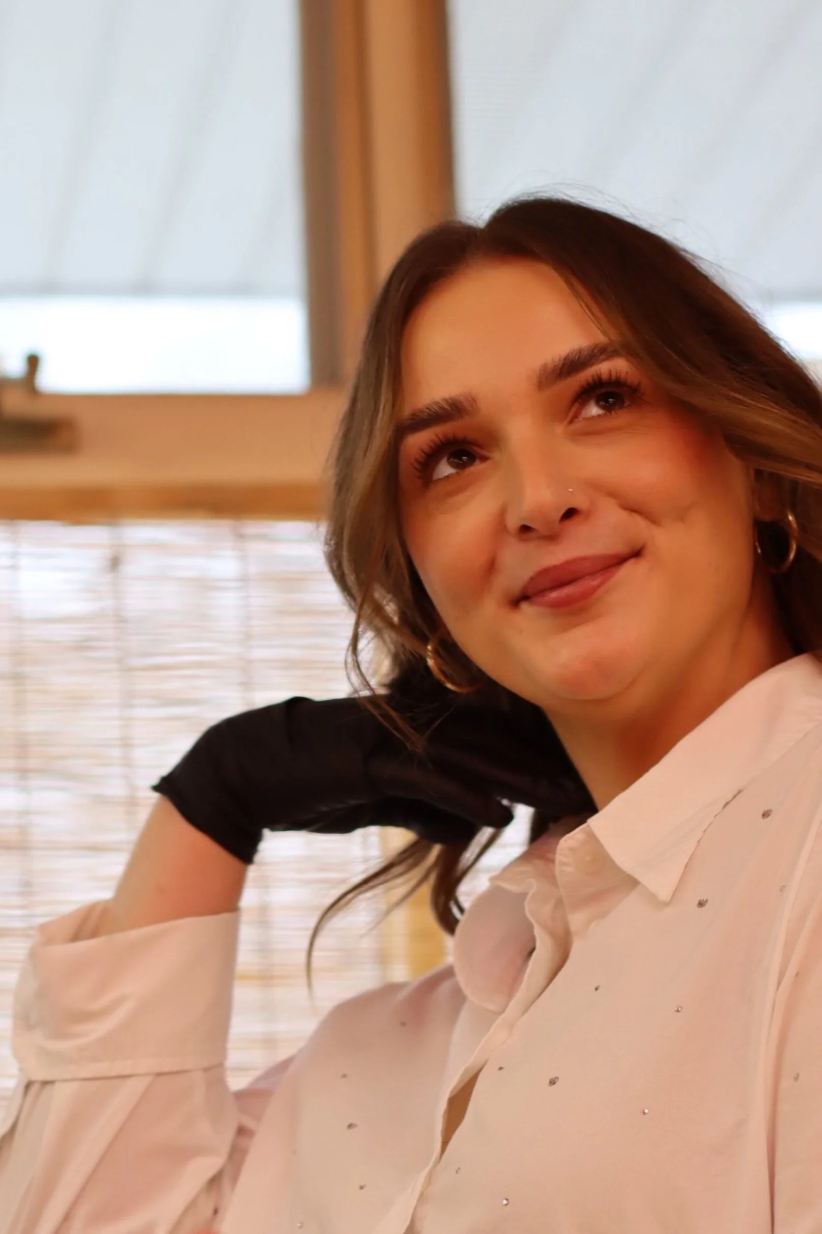 A woman with light brown hair and wearing a light pink blouse, sitting indoors near a window with bamboo window shades, smiling and looking up to her right.