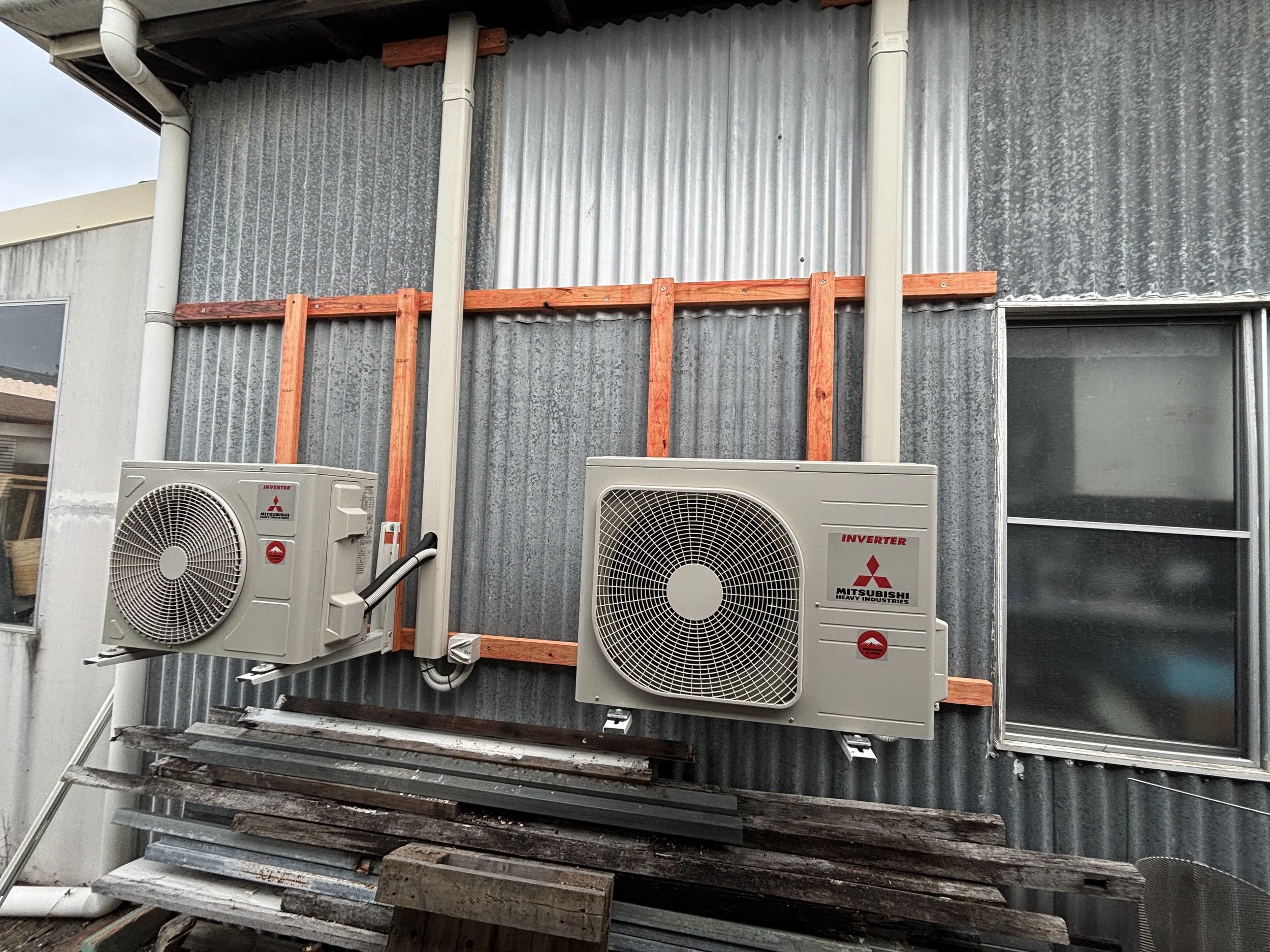 Exterior of a building with two Mitsubishi air conditioning units mounted on a wooden frame constructed of orange wood, and a small window on the right side.