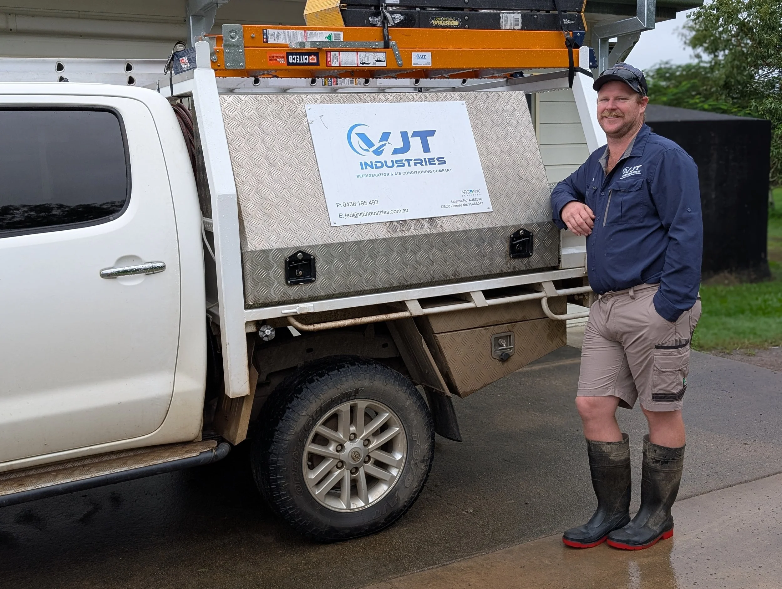 A man standing beside a work truck with a metal toolbox and a ladder on top. The truck has a sign for VJT Industries, a refrigeration and air conditioning company, on the side. The man is wearing a navy blue work shirt, beige shorts, black rubber boots, and a cap, and is smiling at the camera.