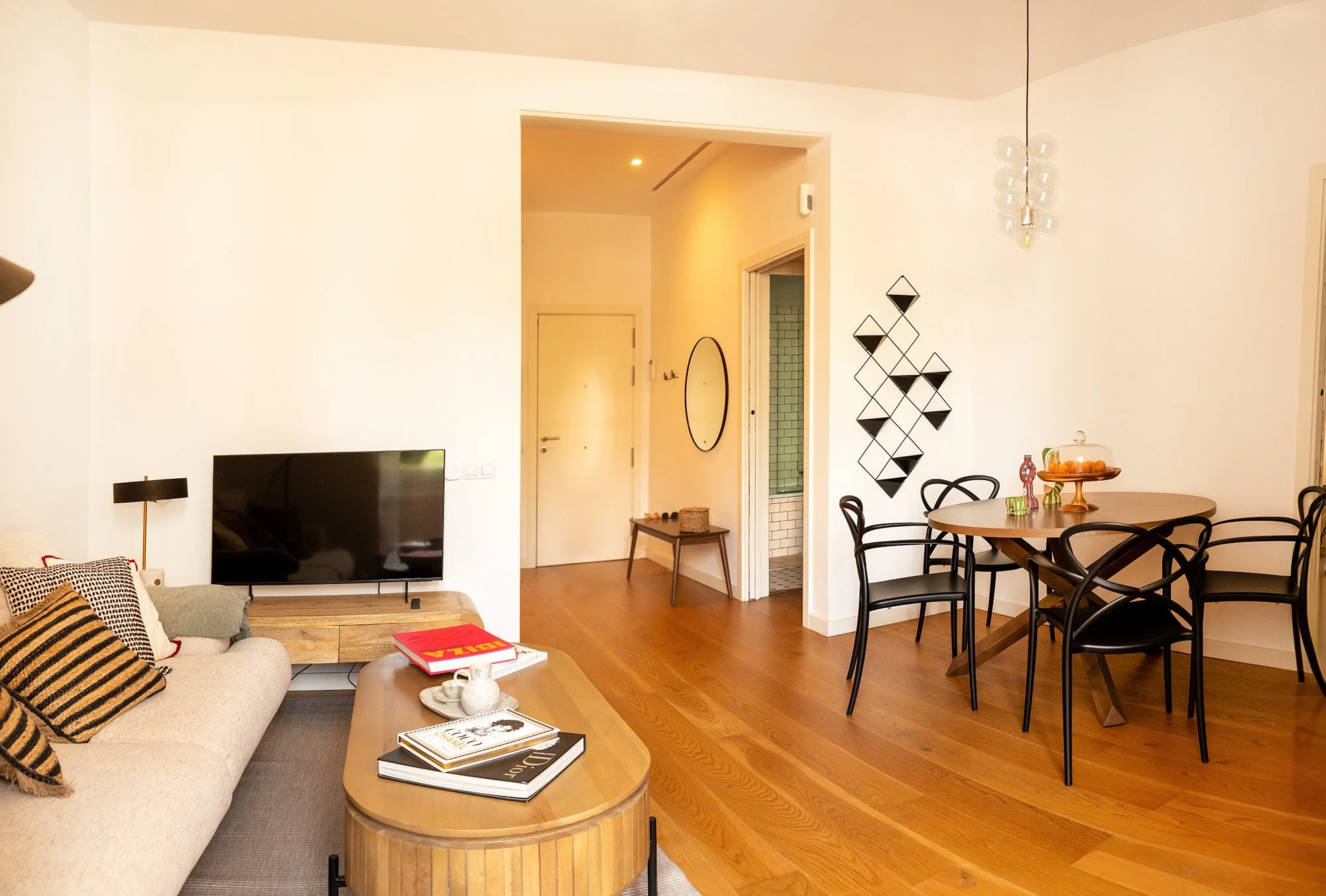 Living room with beige sofa, black TV, wooden coffee table, black dining chairs around a wooden table, white walls, modern black geometric wall decor, side table with accessories, doorway leading to a bathroom with green tiles.