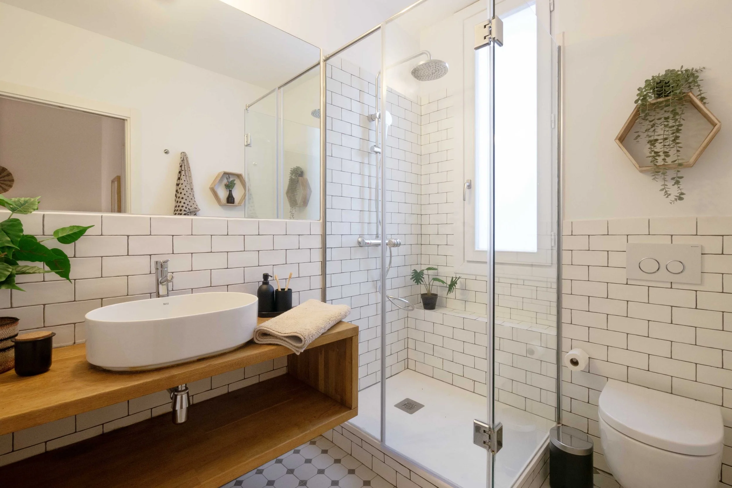 Modern bathroom with white subway tile walls, a wooden vanity with an oval vessel sink, a glass shower enclosure with a window, and decorative wall shelves with plants.