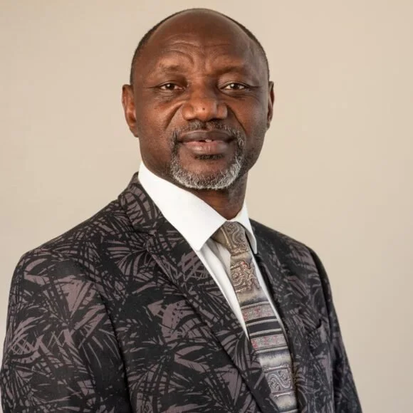 A middle-aged man with a shaved head, beard, and mustache wearing a patterned suit and a striped tie, standing against a plain light-colored background.