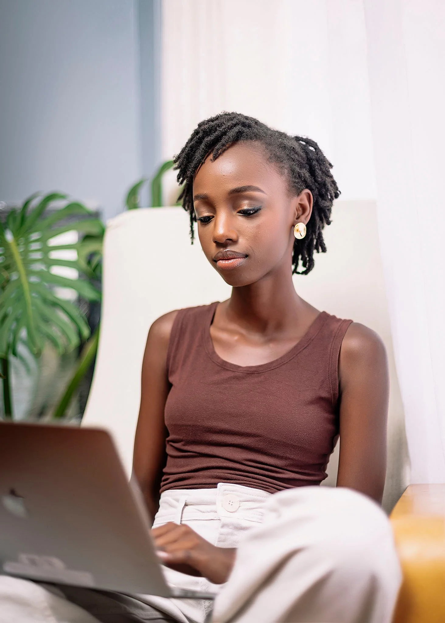 A young Black woman with short dreadlocks, wearing a brown sleeveless top and cream-colored pants, is sitting indoors with a laptop on her lap. She appears focused on her work, with a large green plant and a white curtain in the background.