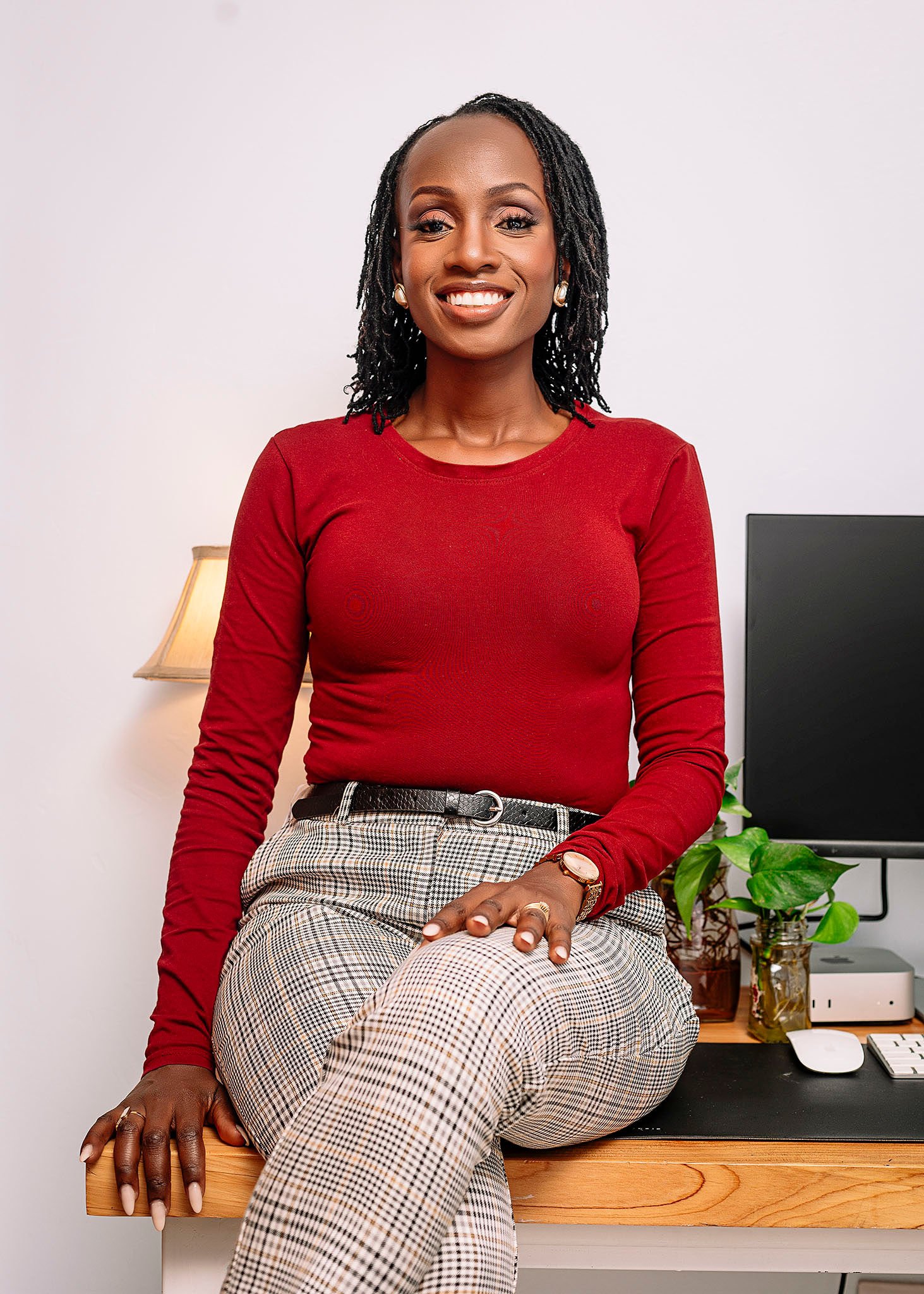 A woman sitting on a wooden desk in an office with a computer and a plant behind her. She is smiling and wearing a red long-sleeve top and plaid pants.