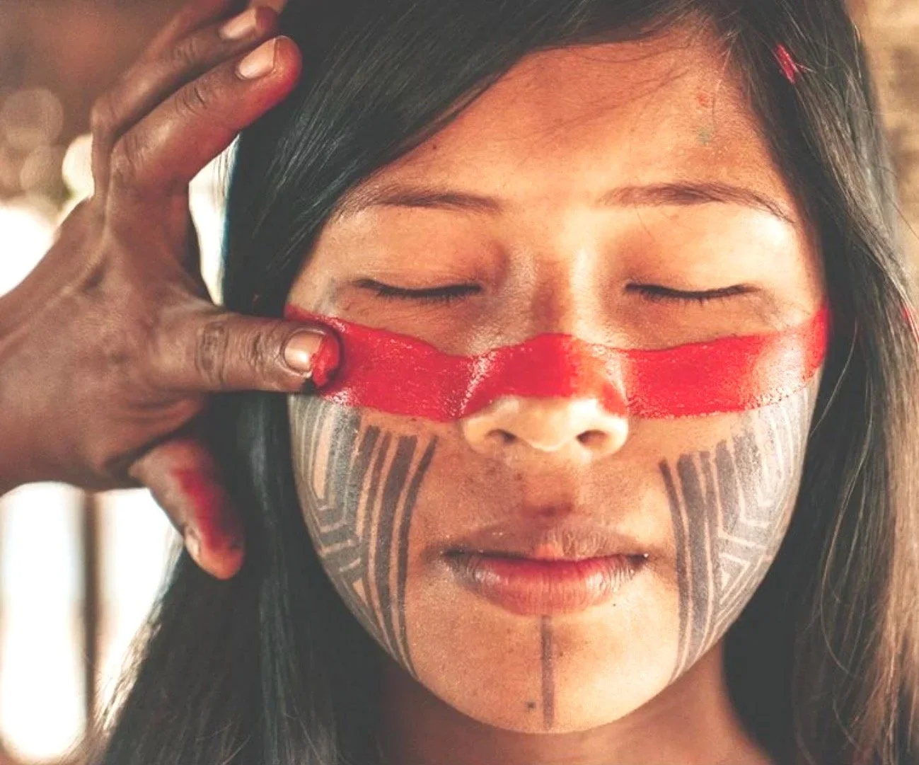 A woman with traditional tribal face paint and tattoos has her face gently touched by a person with darker skin, who appears to be applying or adjusting red body paint across her eyes and nose.