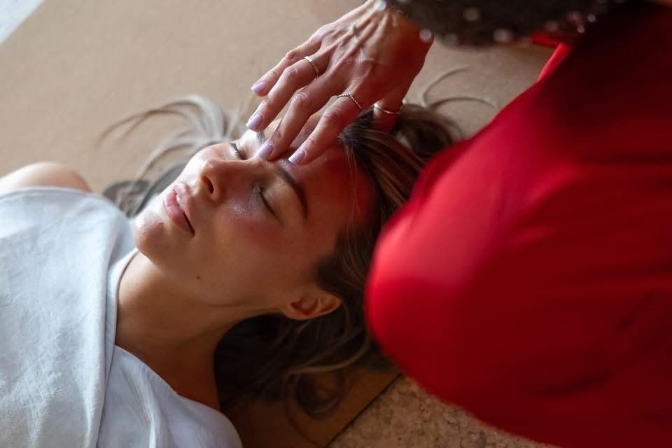 A woman with her eyes closed lying on the floor receiving a forehead massage from another person.
