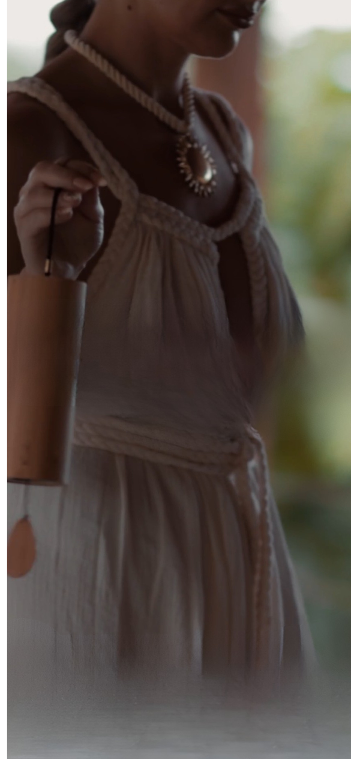 A woman wearing a beige dress and jewelry, holding a cup, with a blurred green background.