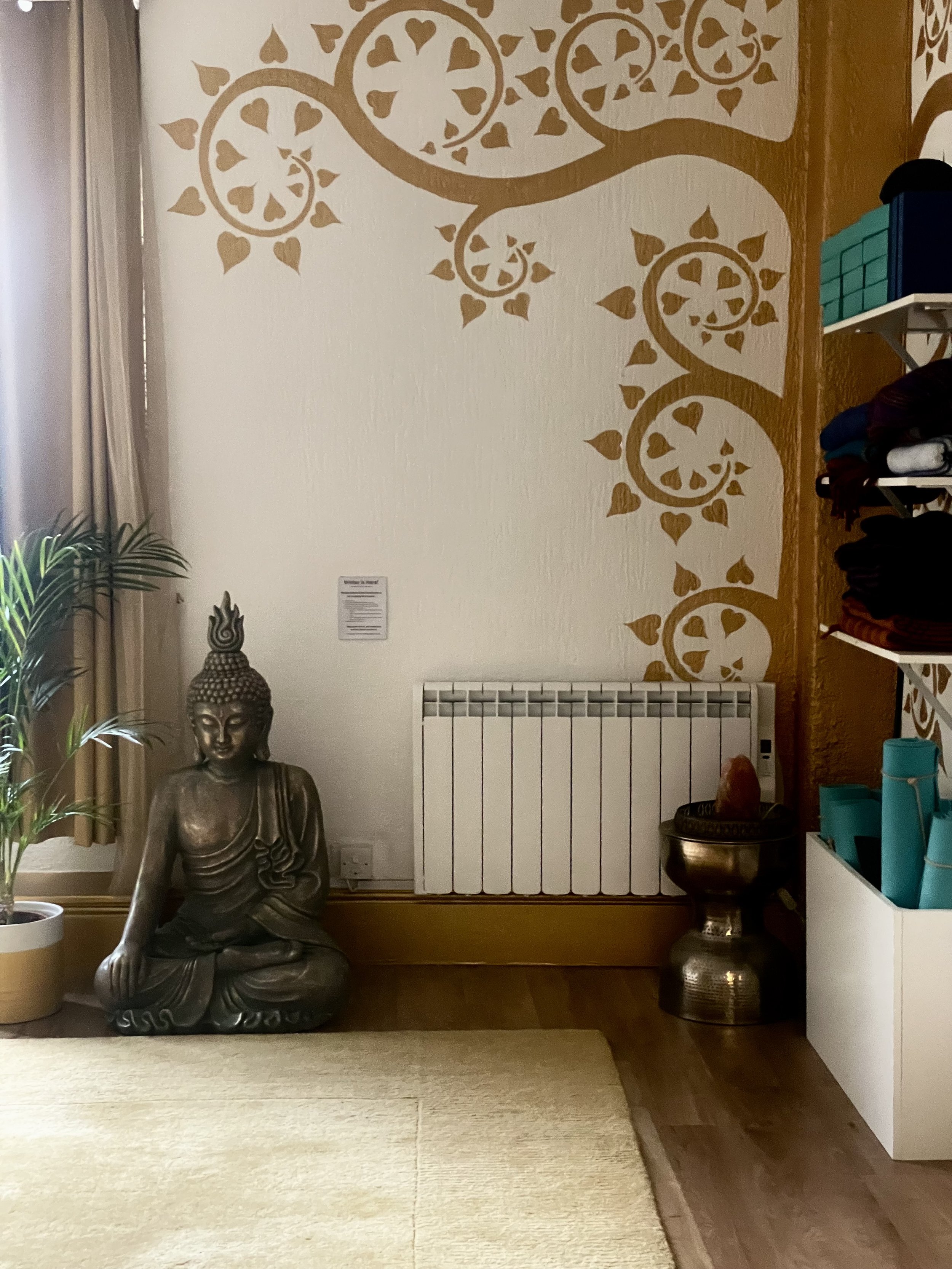 A room with a bronze Buddha statue sitting on the floor, a potted plant to the left, and a white shelf with teal containers on the right. The wall features a decorative gold pattern with swirling circles and heart-shaped elements. The room has a white radiator and various decorations, including a Buddha head sculpture.